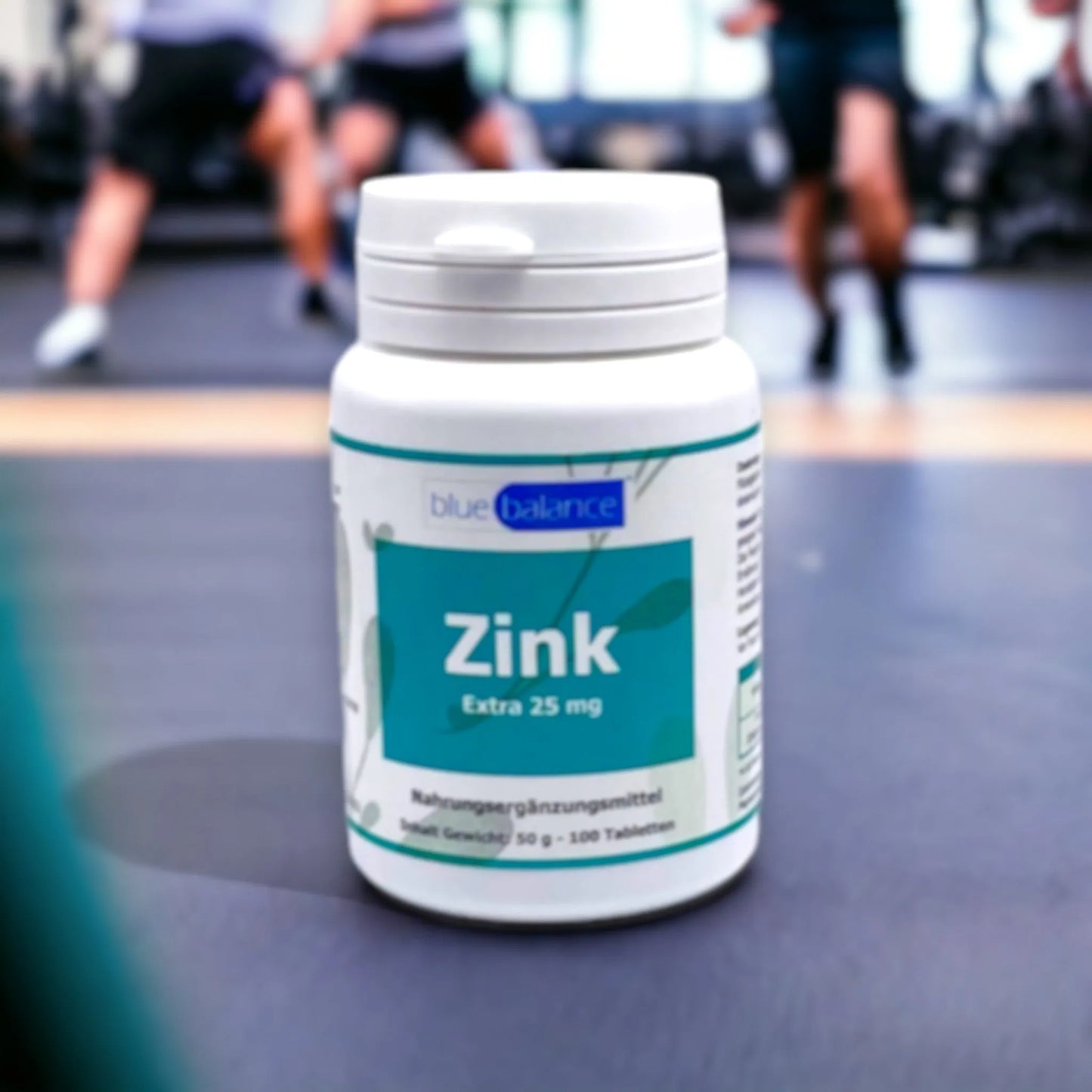 Im Fokus stehen blue balance Zink Tabletten von ETH Meditec vor einem Hintergrund mit verschwommenen Konturen von Menschen, die in einem Fitnessstudio trainieren, symbolisch für Gesundheit und Fitness in Verbindung mit hochwertiger Nahrungsergänzung.